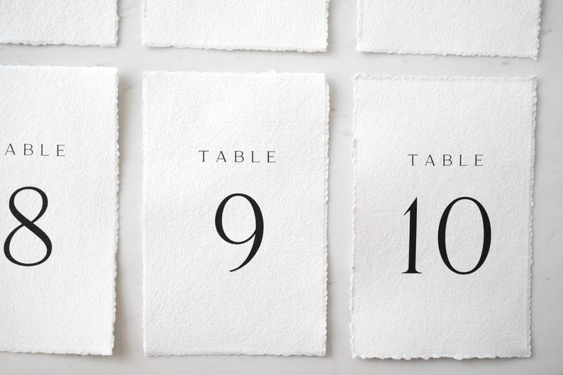 Op de afbeelding: Witte rechthoekige tafelnummerkaarten met gescheurde randen. Het woord "TABLE" staat bovenaan elke kaart in een dun, zwart lettertype. Hieronder staan de grote zwarte cijfers 8, 9 en 10.