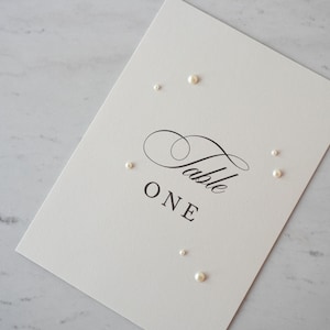 May include: A cream-colored table number card with the word "Table" in elegant script and "ONE" in a simple font. Small, round pearls are scattered around the card, adding a touch of elegance.