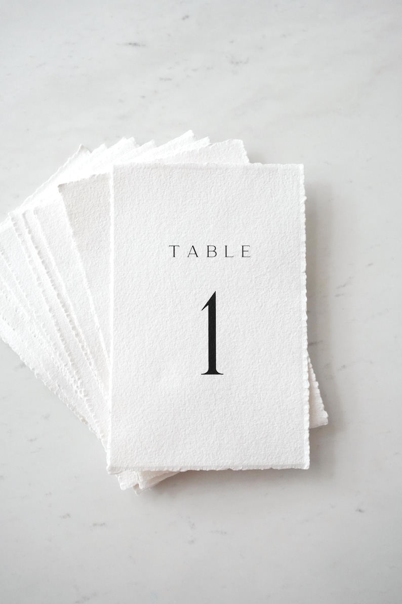 Op de afbeelding: Een stapel witte tafelnummerkaarten met een getextureerde afwerking. De bovenste kaart toont het woord "TABLE" boven het nummer "1" in een vet, zwart lettertype. De kaarten hebben een gescheurde rand, wat ze een rustieke uitstraling geeft.