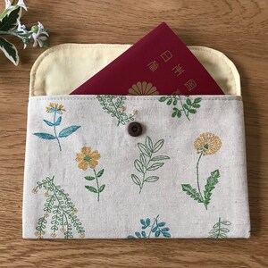 May include: A white linen passport holder with a floral pattern in shades of green and yellow. The holder has a button closure and a red passport is partially visible inside.