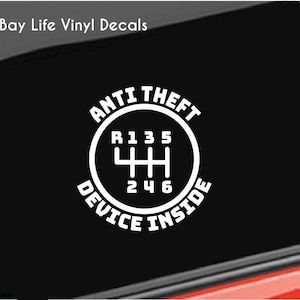 May include: White vinyl decal with the text "ANTI THEFT DEVICE INSIDE" in a circular design, featuring a gear shift pattern. The decal is applied to a car window, with the Bay Life Vinyl Decals logo in the upper left corner.