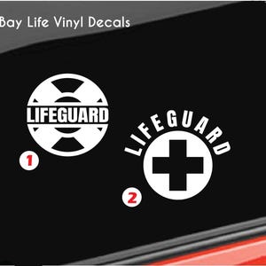 May include: Two white vinyl decals on a black background. Design 1 features a circular life preserver icon with the text LIFEGUARD. Design 2 features a circular border with a central cross and the text LIFEGUARD arched above. Bay Life Vinyl Decals.