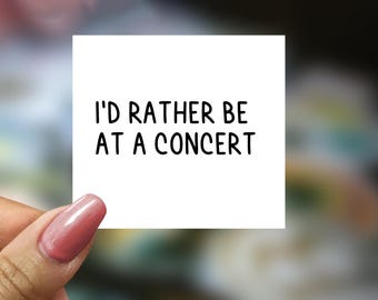 May include: A white square card with the text "I'D RATHER BE AT A CONCERT" in black, held by a hand with a pink nail. The background is blurred with shades of green and white.