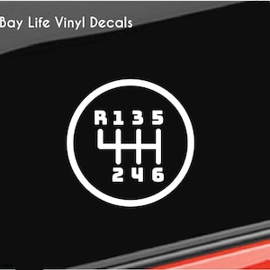 May include: White vinyl decal of a car's manual transmission gear shift pattern, with R, 1, 2, 3, 4, 5, and 6 clearly labeled. The decal is set against a black background, with the text "Bay Life Vinyl Decals" in the upper left corner.