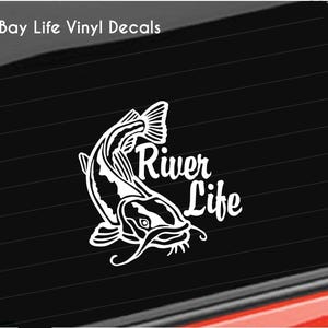 May include: White vinyl decal featuring a stylized catfish leaping upwards. The text "River Life" is written in a decorative font next to the fish. The decal is designed to be applied to a car window or similar surface.