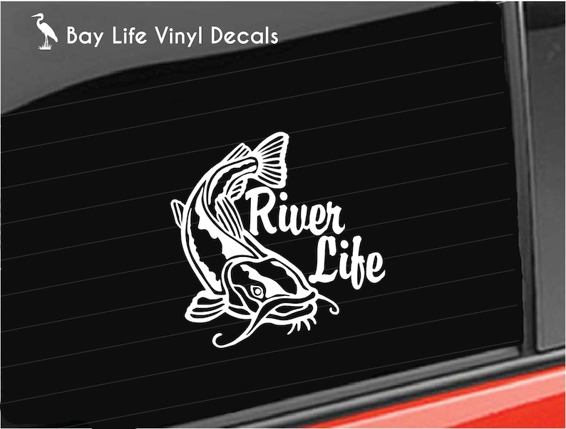May include: White vinyl decal featuring a stylized catfish leaping upwards. The text "River Life" is written in a decorative font next to the fish. The decal is designed to be applied to a car window or similar surface.