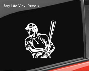 May include: A white vinyl decal featuring a baseball player holding a bat, positioned on a black surface. The text "Bay Life Vinyl Decals" is printed in white in the upper left corner. The design shows the player in a batting stance.
