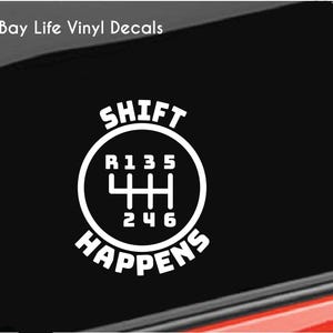 May include: A white vinyl decal applied to a black car window. The design features a circular manual transmission gear shift pattern with the numbers R, 1, 3, 5 on top and 2, 4, 6 on the bottom. The text SHIFT HAPPENS is arched around the circle.