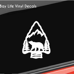 May include: White vinyl decal featuring a bear silhouette walking in front of mountains and trees, inside an arrowhead shape. The decal is on a black background. The text "Bay Life Vinyl Decals" is in the upper left corner.