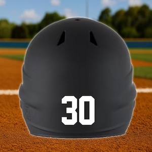 May include: A black baseball helmet with the number 30 in white on the back. The helmet is sitting on a brown surface, with a baseball field in the background.