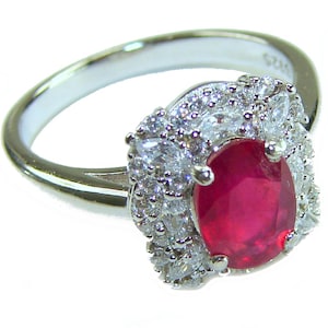 May include: A silver ring featuring a large, oval-cut ruby gemstone surrounded by a halo of smaller, clear gemstones. The ring's band is smooth and polished, and the gemstones sparkle brightly against a white background.