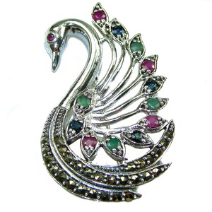 May include: A silver swan brooch with a curved neck and a detailed tail. The brooch features a variety of colourful gemstones, including red, green, and blue stones. The swan's eye is a single red gemstone.