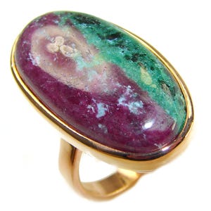 Puede incluir: Un anillo de oro con una gran gema ovalada. La gema presenta una mezcla de tonos rojo intenso, verde azulado y verde, con una superficie pulida. La banda y el engaste del anillo son dorados, creando un contraste llamativo.