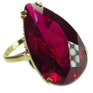 Op de afbeelding: Een gouden ring met een grote, facetgeslepen, druppelvormige, dieprode edelsteen. De ring heeft een gedetailleerde gouden band en decoratieve accenten rond de edelsteen setting. De slijping van de edelsteen creëert meerdere reflecterende oppervlakken.