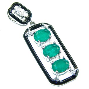 Puede incluir: Colgante rectangular con borde negro y tres esmeraldas ovaladas. El colgante tiene un engaste plateado y una pequeña sección superior rectangular con una piedra transparente. Las esmeraldas son de color verde vibrante.