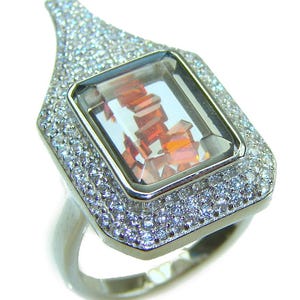 May include: A silver ring featuring a rectangular, orange-hued gemstone at its center. The gemstone is surrounded by a frame of smaller, clear stones. The ring's band is a simple, polished silver.