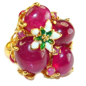 May include: A gold-toned ring featuring large, oval-shaped, deep red gemstones clustered together. The ring is accented with a white and green floral design and smaller red gemstones. The ring has a unique, ornate design.