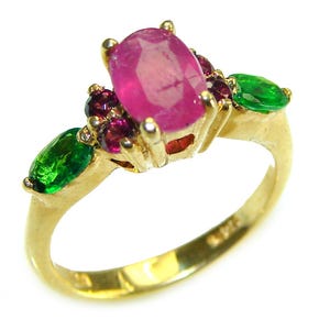 Op de afbeelding: Een gouden ring met een grote ovale robijn als middensteen, omringd door kleinere rode en groene edelstenen. De ring heeft een klassiek ontwerp met een gepolijste afwerking, die de levendige kleuren van de edelstenen benadrukt.