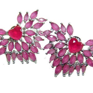 May include: A pair of silver-toned earrings featuring a heart-shaped, deep pink center stone surrounded by numerous smaller, oval-shaped pink stones. The earrings have a floral or starburst design.