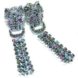 May include: A pair of silver-toned drop earrings. Each earring features a jaguar head with red gemstone eyes and a dangling chain of colorful gemstones. The earrings are detailed with small, dark-colored stones.