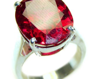 May include: A silver ring featuring a large, faceted, oval-cut, deep red gemstone. The gemstone is held in place by four silver prongs. The ring band is a simple, polished silver design. The gemstone's color is a vibrant, saturated red.
