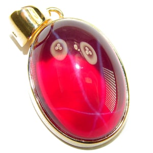 Puede incluir: Colgante ovalado con una gema roja vibrante. La piedra tiene vetas blancas y moradas e inclusiones blancas circulares. El colgante está engarzado en un marco plateado con una fianza dorada.