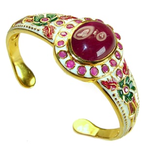 May include: Gold-tone cuff bracelet with a large oval ruby gemstone centre. The bracelet features a white enamel setting with small pink gemstones and floral designs in red, green, and white. The open design allows for easy wear.