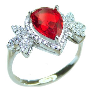 Puede incluir: Un anillo de piedra roja en forma de pera con una banda de plata y un diseño de mariposa a cada lado. El anillo está engastado con piedras transparentes más pequeñas.