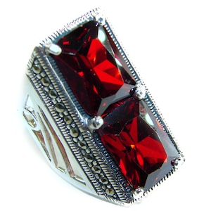 May include: A silver ring with a large, rectangular, deep red gemstone. The ring has a detailed setting with small, round, dark accents and a decorative band. The gemstone is faceted.