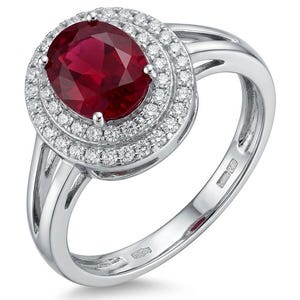 May include: A silver-toned ring featuring a large, oval-cut deep red gemstone at the center. The stone is surrounded by a double halo of small, clear, round-cut stones. The band is polished metal with a split-shank design and a visible hallmark.