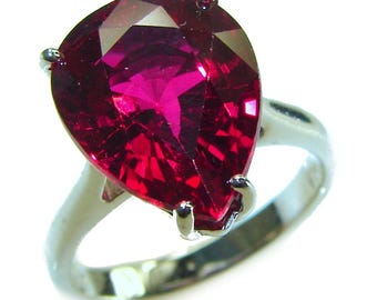 May include: A pear-shaped, faceted, deep red gemstone ring. The gemstone is set in a silver-colored band with four prongs. The ring's design is simple, highlighting the vibrant color and cut of the stone.