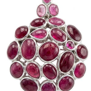 May include: A circular silver brooch featuring numerous cabochon-cut magenta and deep pink gemstones arranged in a cluster. The stones are set in polished silver bezels, creating a dense, rounded composition with a small triangular bail at the top.