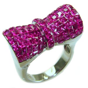 May include: A silver ring featuring a large, bow-shaped design encrusted with numerous square-cut, vibrant pink gemstones. The ring's band is a polished silver, and the gemstones are closely set, creating a dazzling display.
