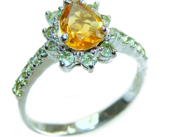 May include: A silver ring featuring a pear-shaped, golden-yellow gemstone at its center, surrounded by smaller, light green gemstones. The ring's band is also adorned with small, light green gemstones, creating a sparkling, elegant design.