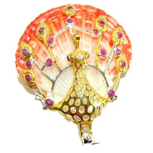 May include: A decorative peacock brooch with gold and silver accents, adorned with small red gemstones. The peacock design is set against a natural seashell with orange and white hues, creating a unique jewelry piece.
