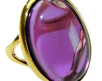 May include: A gold-toned ring featuring a large, oval-shaped, translucent purple stone. The stone is set within a gold bezel, and the ring's band is also gold. The ring is designed to be worn on a finger.