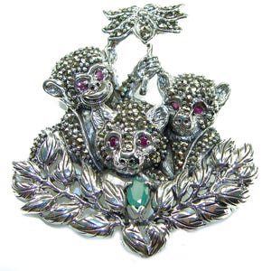 Op de afbeelding: Zilverkleurige broche met drie apenfiguren met robijnen ogen en markasietdetails. De apen zijn omgeven door bladeren en houden een decoratief element vast. Een smaragdgroene steen is aan de basis geplaatst.