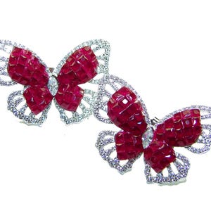 May include: Two butterfly-shaped brooches. Each brooch features a silver-colored, openwork frame encrusted with small, clear stones. The wings are filled with faceted, deep red stones, and a clear, elongated stone is at the center.