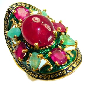 May include: An oval-shaped ring with a gold band, featuring a large, round, red gemstone at the center, surrounded by smaller red and green gemstones. The ring has a green enamel base with gold accents.