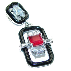 Puede incluir: Un colgante de plata con una piedra preciosa roja rodeada de piedras preciosas transparentes. El colgante está engastado en un marco de esmalte negro.