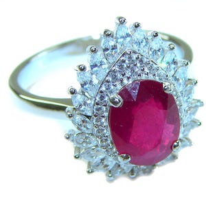 May include: A silver ring featuring a large, oval-cut ruby center stone. The ruby is surrounded by a halo of smaller, clear, marquise-cut and round-cut gemstones. The ring's band is a simple, polished silver.