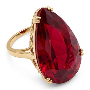May include: A gold ring featuring a large, pear-shaped, faceted deep red gemstone. The setting includes intricate, openwork metal detailing along the sides of the band, which supports the prominent, vertically oriented, teardrop-shaped stone.