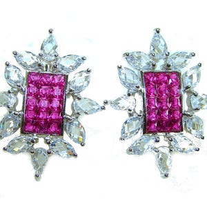 May include: A pair of silver-toned stud earrings. Each earring features a rectangular center of small, pink gemstones, surrounded by clear, teardrop-shaped stones. The design is symmetrical and elegant, with a floral-like arrangement.