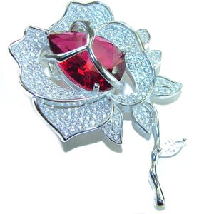 Puede incluir: Broche de rosa plateada con una gran gema roja en forma de pera en el centro. Los pétalos y las hojas están incrustados con pequeños cristales transparentes. El broche tiene un tallo y una sola hoja.