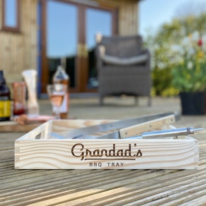 May include: A light-colored wooden BBQ tray with the words "Grandad's BBQ TRAY" engraved on the front. A carving knife and fork rest inside the tray. The tray is on a wooden deck, with a blurred outdoor scene in the background.
