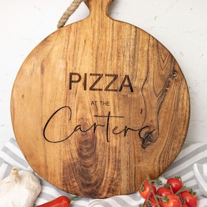 Könnte beinhalten: Rundes Pizza-Servierbrett aus Holz mit Griff und Seildetail. Der Text "PIZZA at the Carter's" ist auf das Brett graviert. Frische Tomaten, Knoblauch und eine rote Paprika umgeben das Brett.