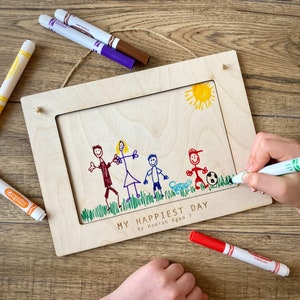 May include: A wooden picture frame with a child's drawing of a family, sun, and soccer ball. The drawing is in red, blue, green, and yellow marker. The text "MY HAPPIEST DAY by Hamish Aged 7" is at the bottom.