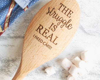 Puede incluir: Una cuchara de madera con el texto "The struggle is real I need cake!" grabado en ella. La cuchara es de color marrón claro y tiene un acabado de madera natural.