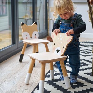 May include: Two wooden children's chairs with animal faces. The chairs are painted in natural wood tones with white and black accents. A child is sitting on one of the chairs, which has a deer face.