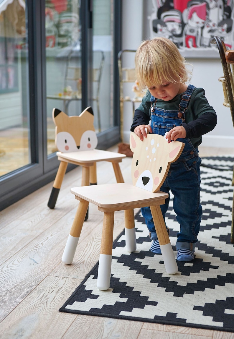 May include: Two wooden children's chairs with animal faces. The chairs are painted in natural wood tones with white and black accents. A child is sitting on one of the chairs, which has a deer face.